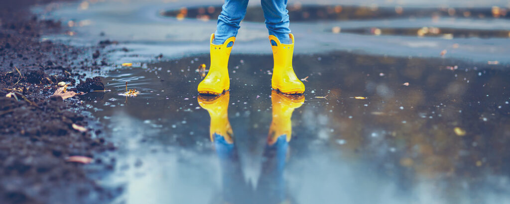 un enfant avec des bottes et un ciré jaune, les pieds dans l'eau